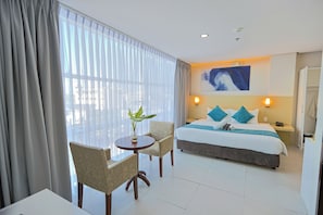 Quarto de hotel com cama, duas cadeiras, uma mesinha com uma planta e uma janela grande com cortinas.