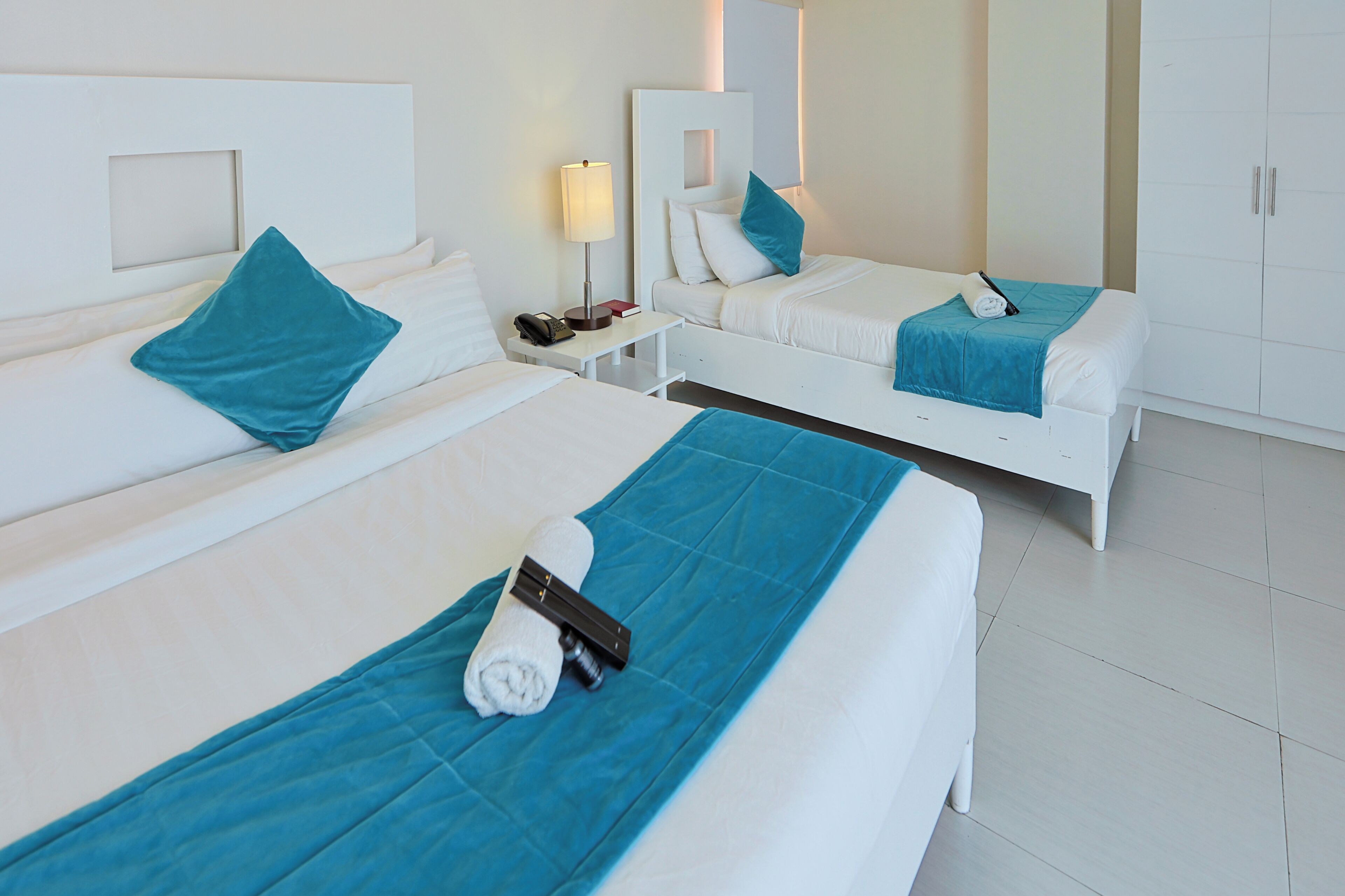A hotel room with two beds, each with a blue bedspread, white headboards, and a nightstand with a lamp and a phone.