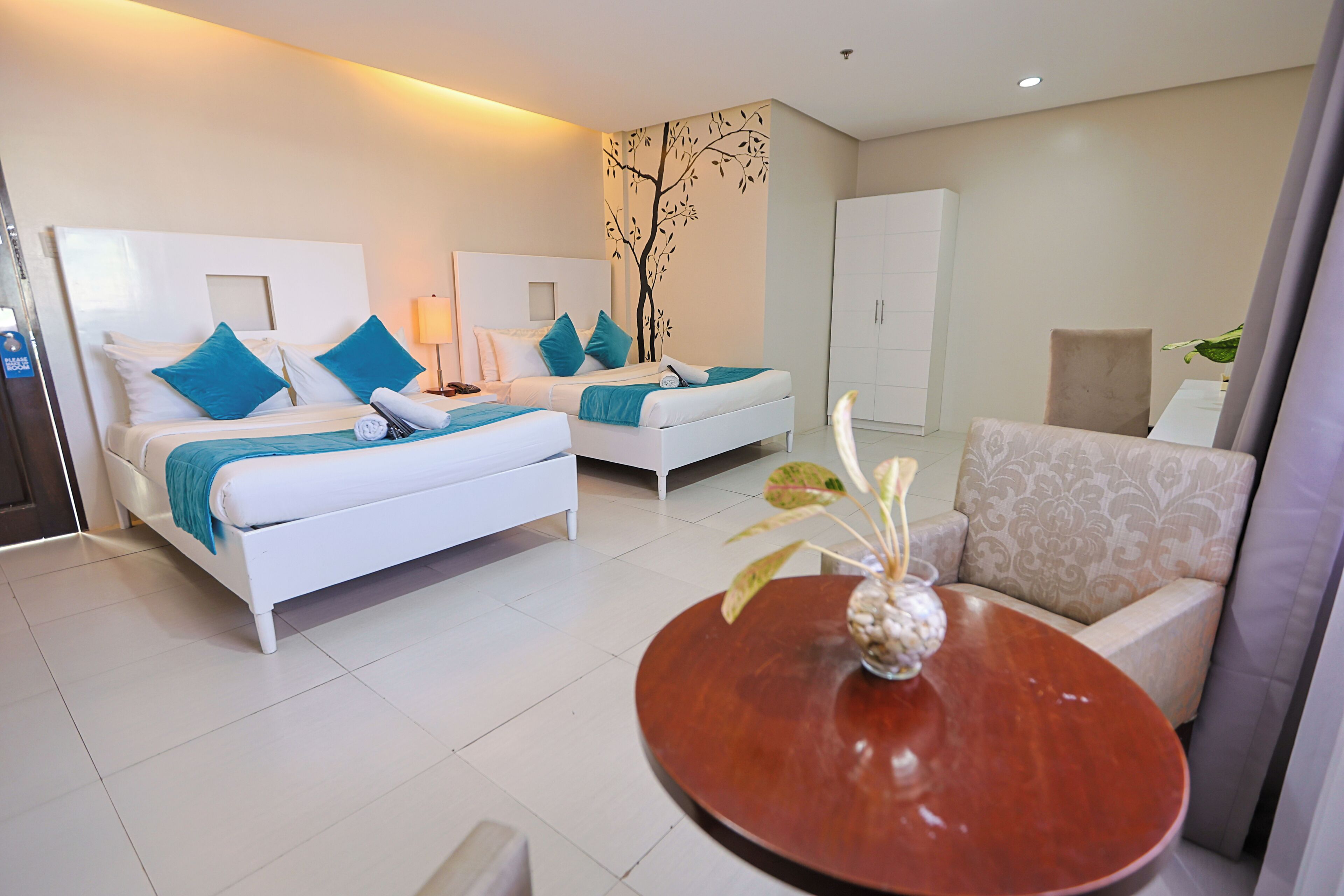 A hotel room with two beds, a sofa, a round table, and a wall-mounted tree decal.