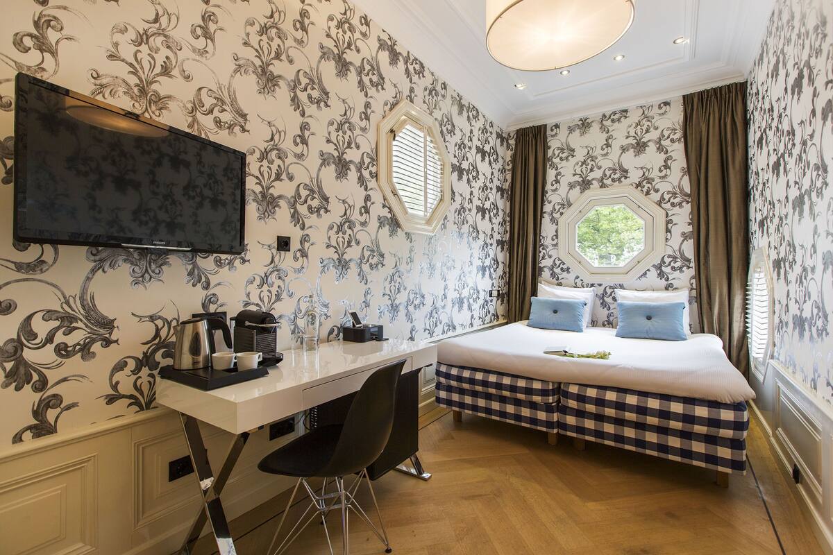 Une chambre d’hôtel comprenant un lit, un bureau avec une chaise, une télévision et une fenêtre avec des rideaux.