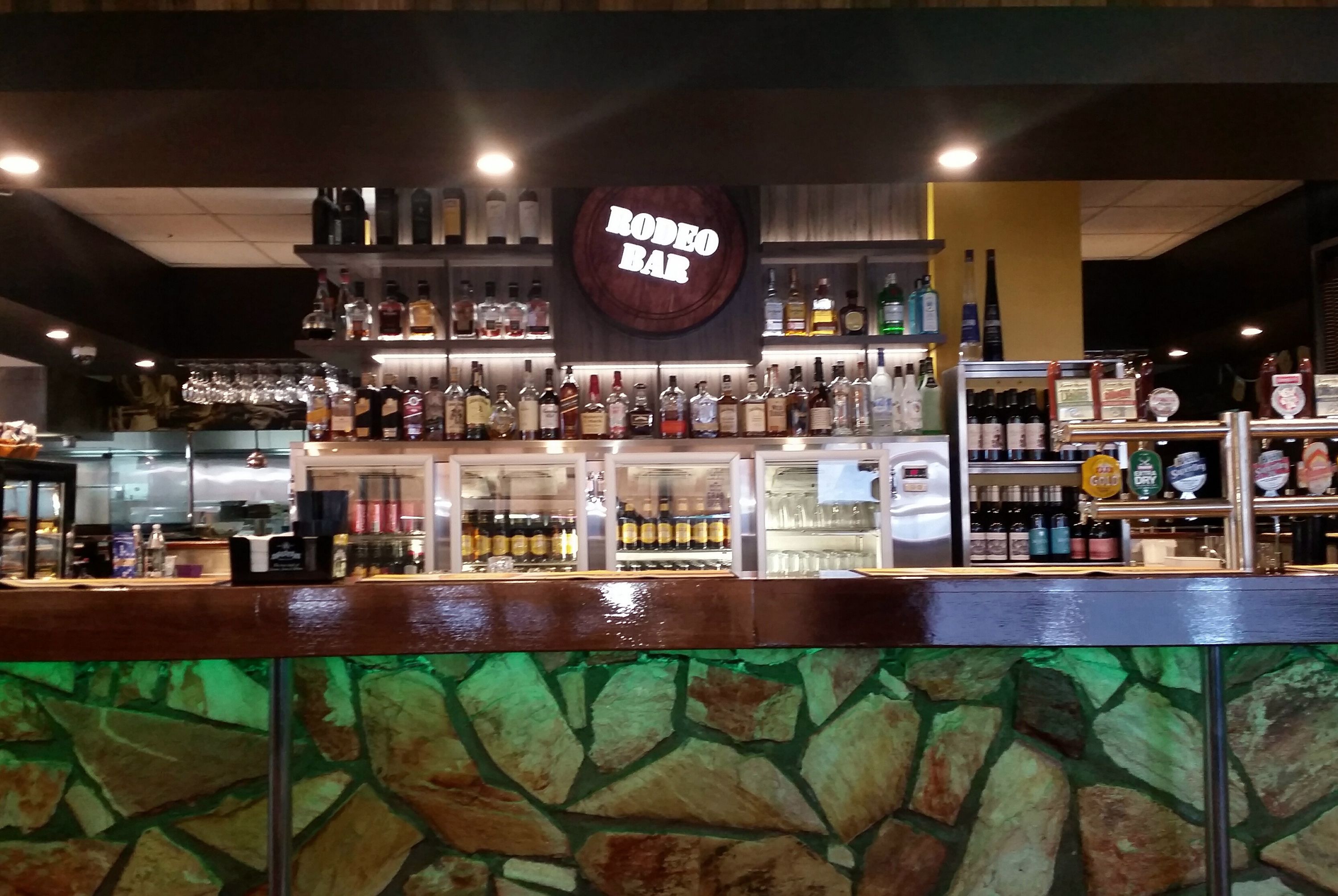 A bar with a 'RODEO BAR' sign, shelves of liquor bottles, and a stone wall in the foreground.