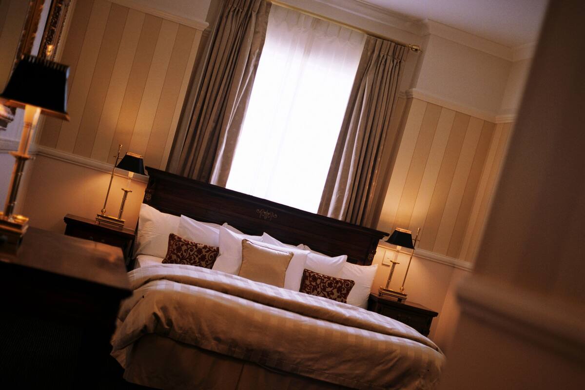 Una cama bien hecha con cabecera de madera, dos mesitas de noche con lámparas y una ventana con cortinas.