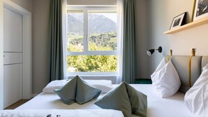 A bedroom with a bed, pillows, a nightstand, a lamp, a shelf with pictures, and a window with a mountain view. - City Hotel Merano (Merano)