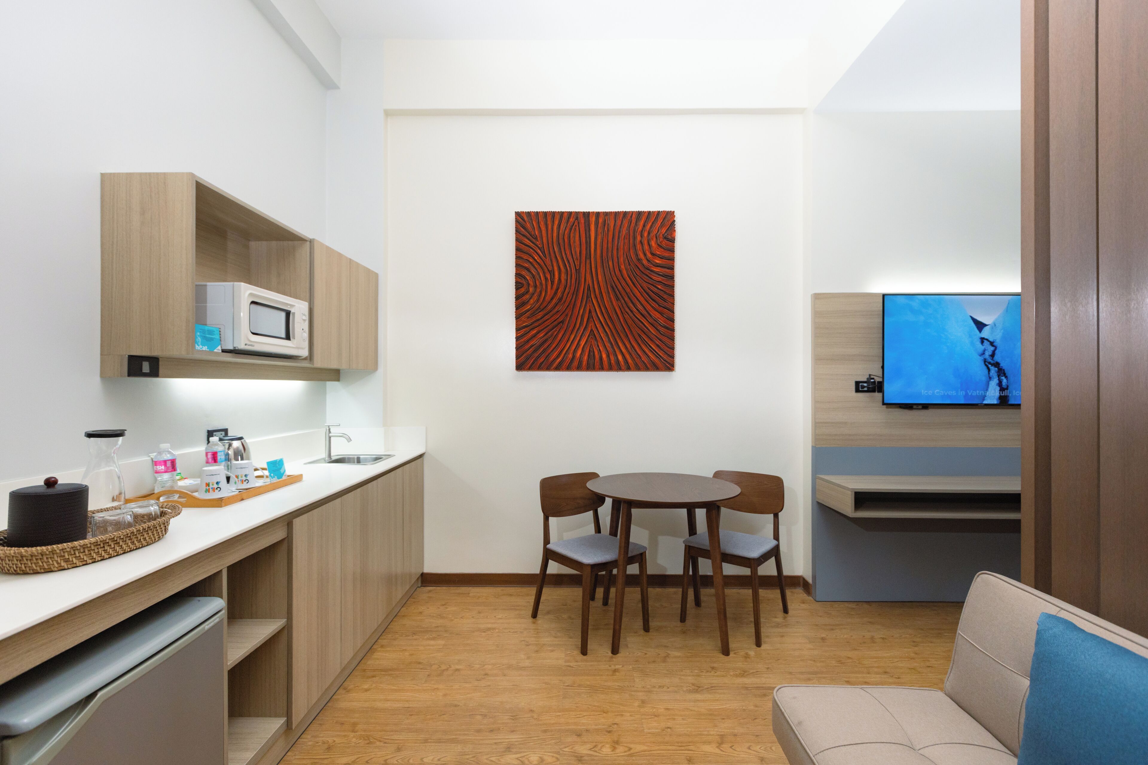 A compact kitchen with a microwave, wooden cabinets, a dining table with chairs, a flat-screen TV, and a sofa.