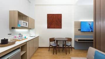 A compact kitchen with a microwave, wooden cabinets, a dining table with chairs, a flat-screen TV, and a sofa.