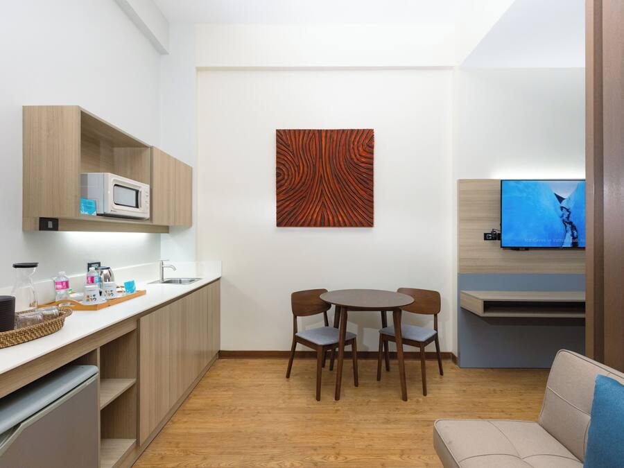 A compact kitchen with a microwave, wooden cabinets, a dining table with chairs, a flat-screen TV, and a sofa.