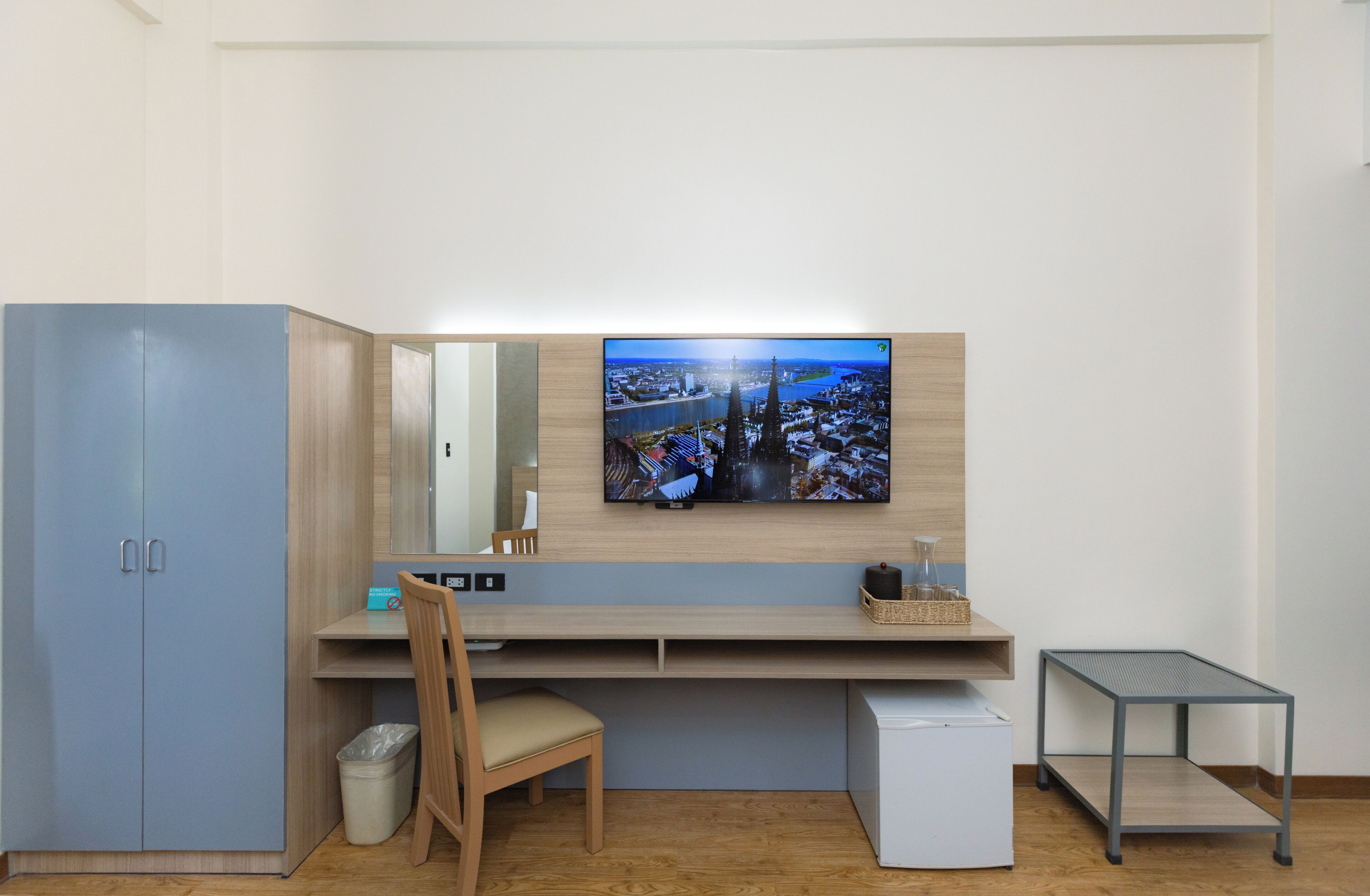 A modern hotel room with a wooden desk, a chair, a small table, a TV mounted on the wall, and a wardrobe.