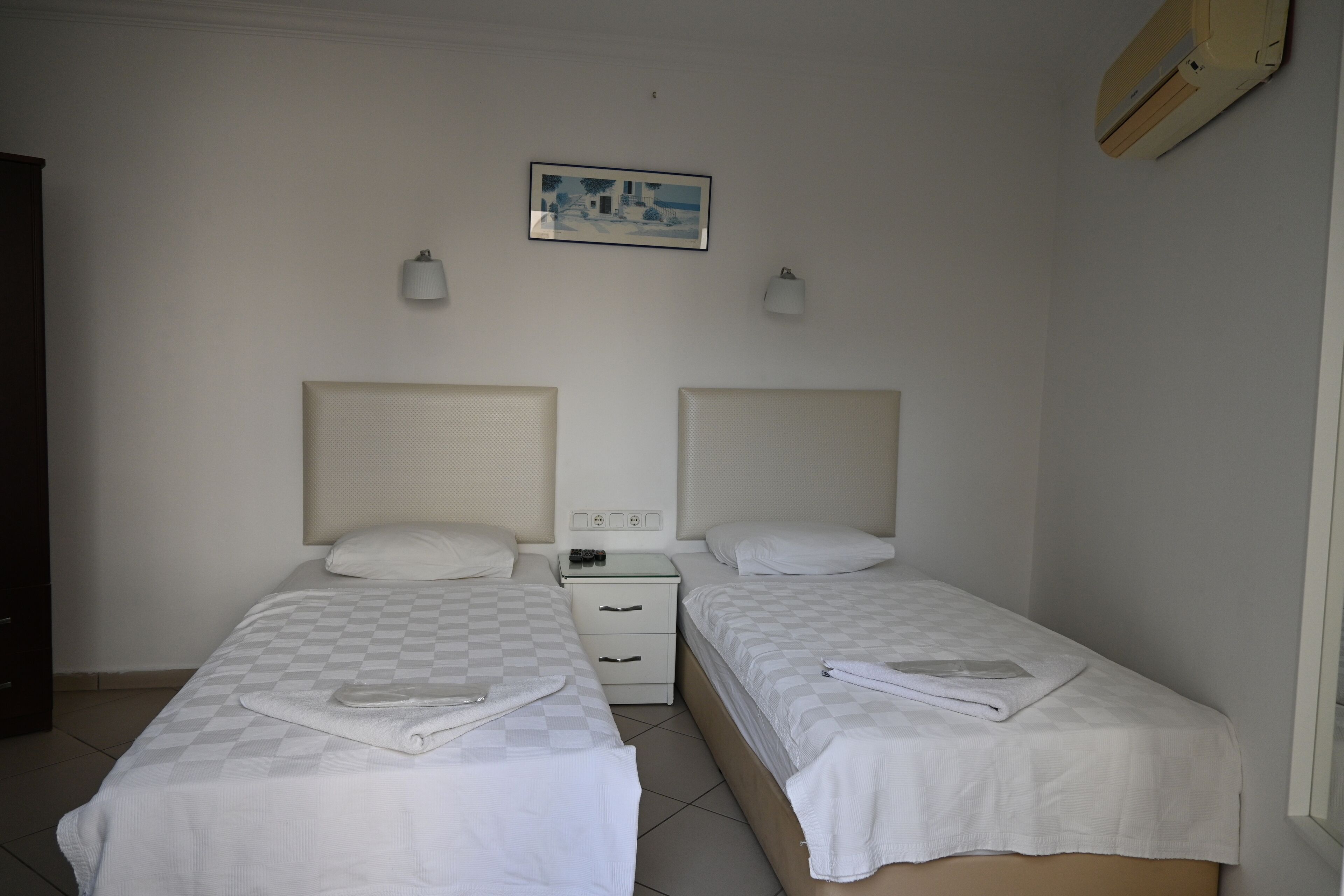 A room with two beds, a small bedside table, a wall-mounted air conditioner, and a framed picture on the wall.