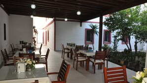 A covered outdoor dining area with wooden tables and chairs, potted plants, and a view of a white building with red shutters. - Melis Pansiyon (Bodrum)
