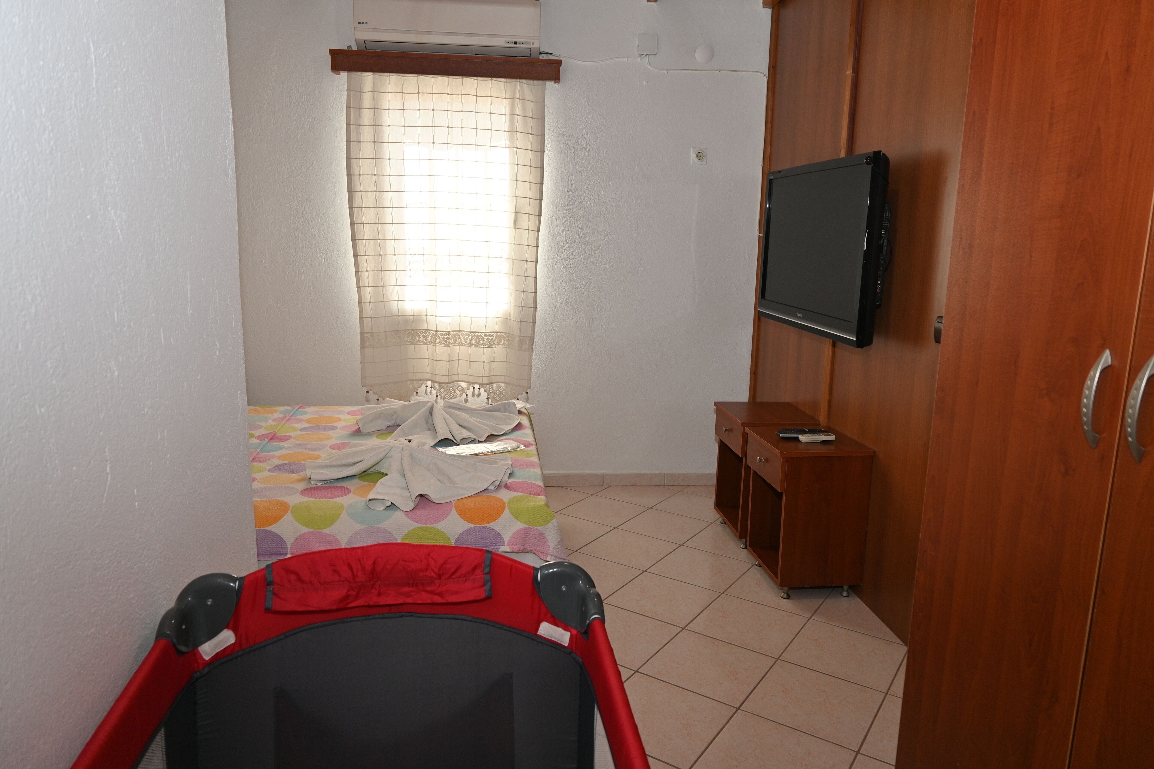 A bedroom with a bed, a TV mounted on the wall, a wooden wardrobe, and a stroller in the foreground.