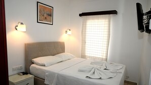A neatly made bed with a headboard, two bedside lamps, a nightstand with drawers, a window with blinds, and a framed picture on the wall. - Melis Pansiyon (Bodrum)