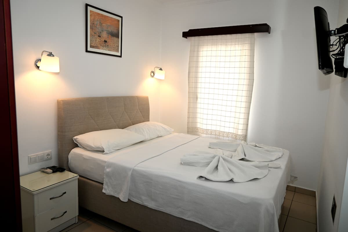 A neatly made bed with a headboard, two bedside lamps, a nightstand with drawers, a window with blinds, and a framed picture on the wall.