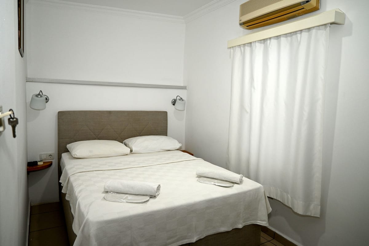 A neatly made bed with white linens and towels, a bedside table with a lamp, and an air conditioning unit above the bed.