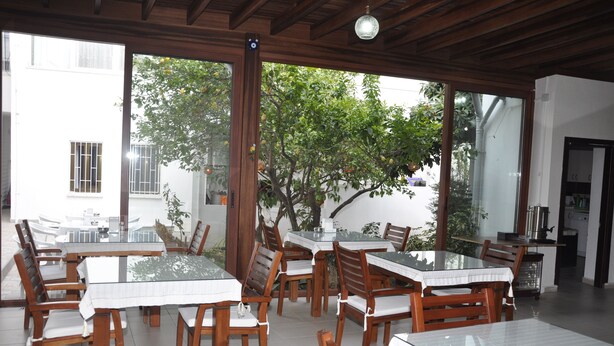 A dining area with multiple tables and chairs, large windows with a view of trees and a building, and a kitchen area visible in the background. - Melis Pansiyon (Bodrum)