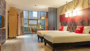 Une chambre d’hôtel avec deux lits, une armoire en bois, une petite table avec une chaise et une grande fenêtre donnant sur la ville.