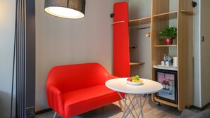Une chambre moderne avec un canapé rouge, une petite table ronde sur laquelle sont disposés des fruits, un mini-réfrigérateur et une bibliothèque agrémentée d’une plante.