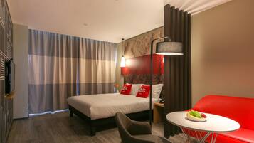Habitación de hotel con una cama, un sofá rojo, una mesita con fruta, una lámpara y un televisor.
