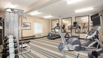 Une salle de sport équipée de matériel d’exercice, de miroirs et d’une fenêtre avec des stores.