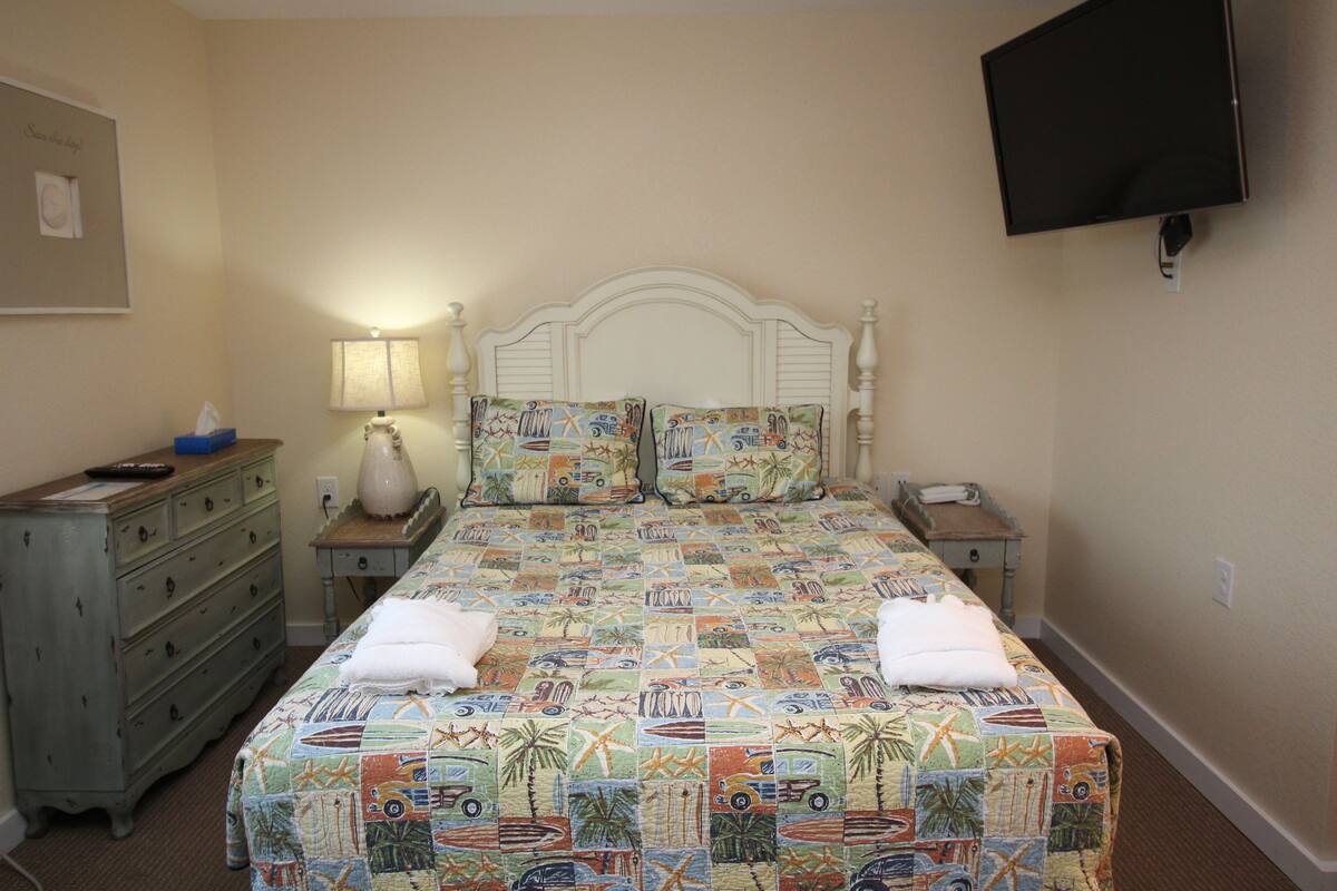 A bedroom with a bed, a dresser, a nightstand, a lamp, and a TV mounted on the wall.