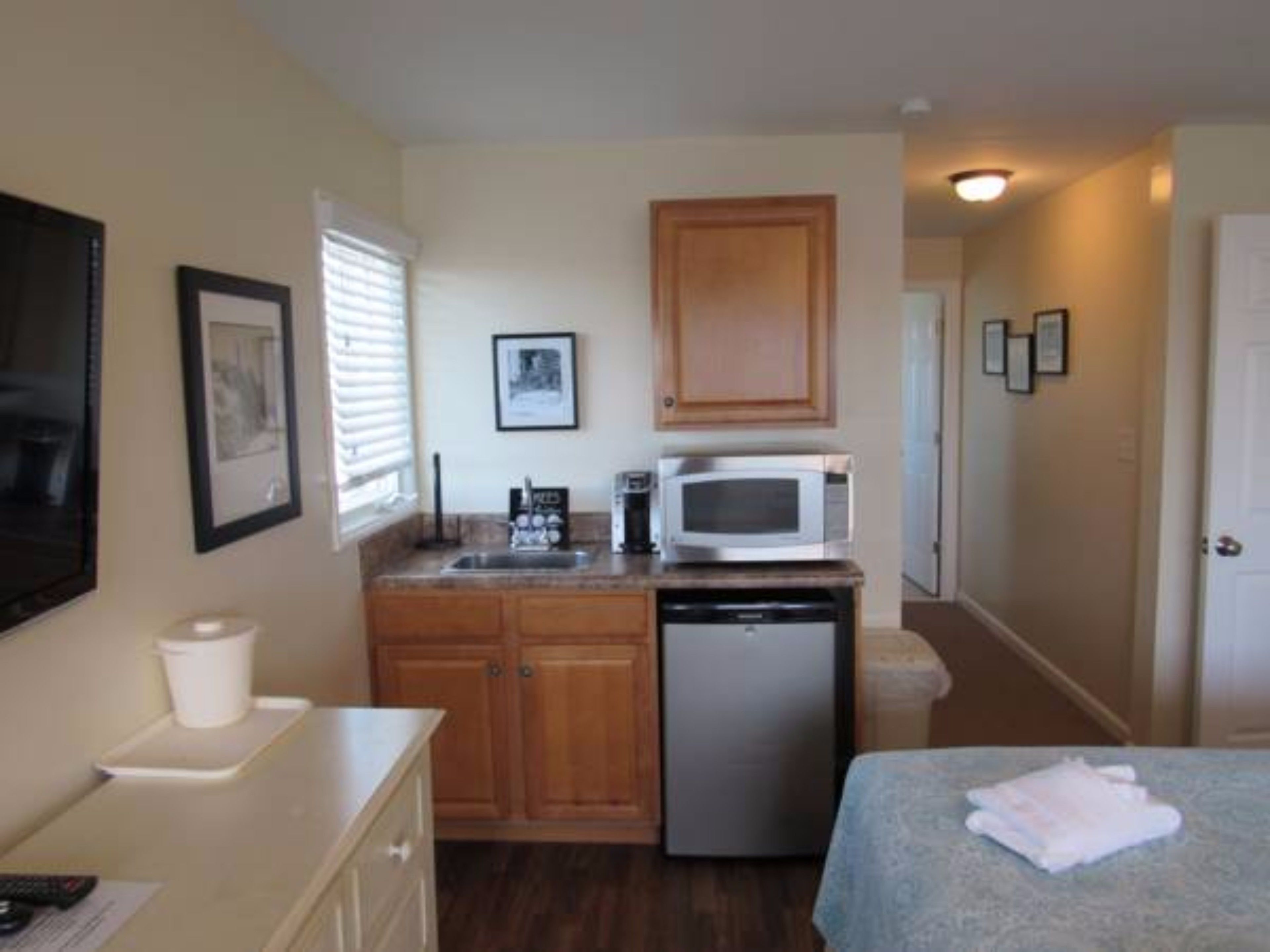A compact kitchenette with a microwave, sink, and small refrigerator. There is a bed with a blanket and a cup on a dresser.
