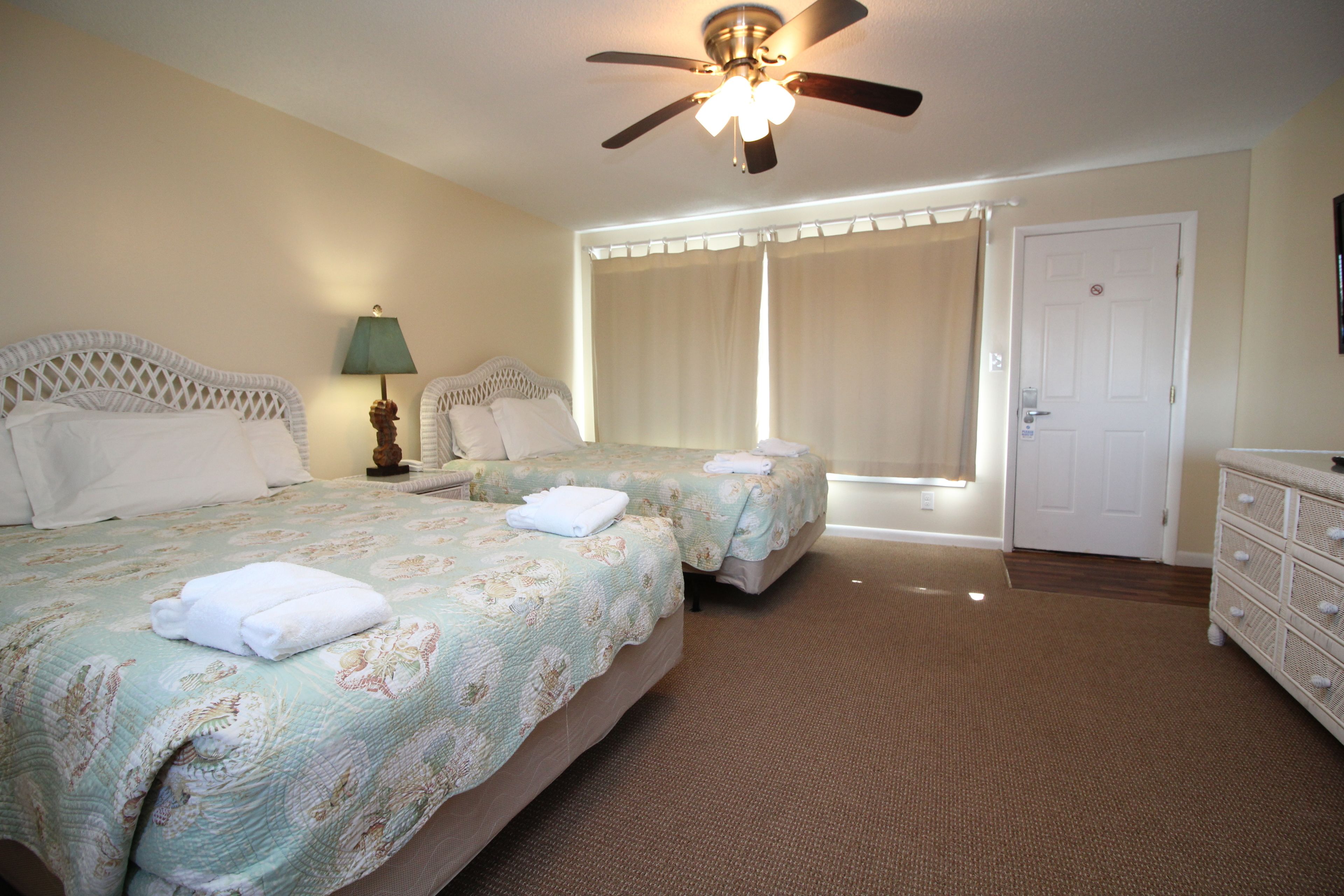 A hotel room with two beds, a ceiling fan, a nightstand with a lamp, a dresser, and a door.