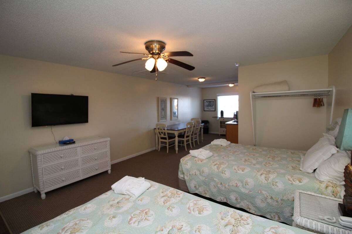A hotel room with two beds, a ceiling fan, a television, a dresser, and a desk with chairs.