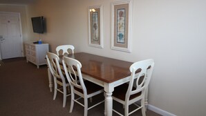 A dining area with a wooden table and four white chairs. There is a television mounted on the wall and a small cabinet with drawers. - Fin 'n Feather Waterside Inn By Kees Vacations (Nags Head)
