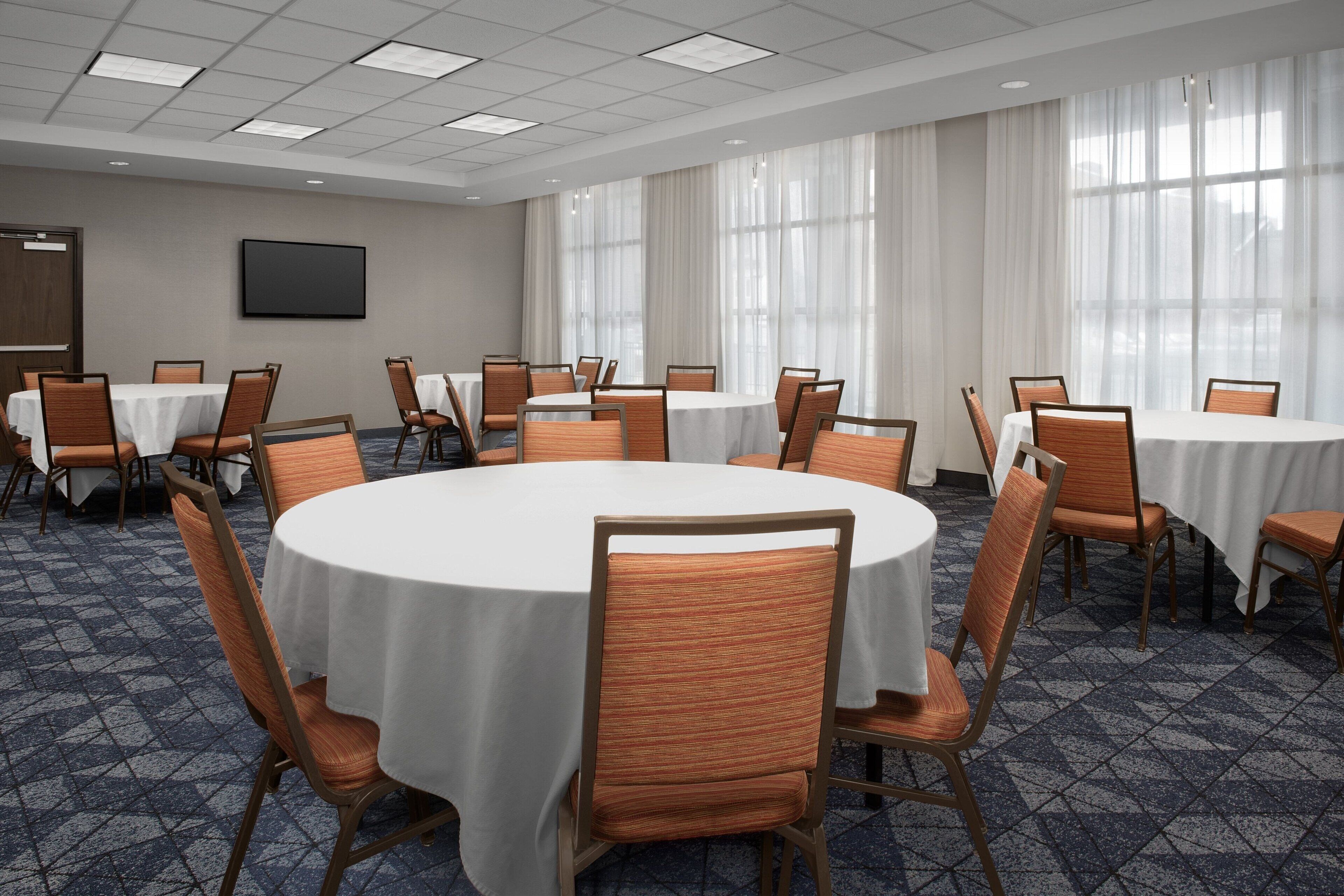 Un salón de conferencias con mesas redondas, sillas, un televisor de pantalla plana y ventanas grandes con cortinas translúcidas.