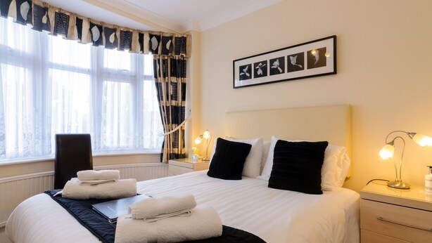 A neatly made bed with white and black linens, a bedside table with a lamp, and a window with sheer curtains. - The Gables Guest House (Lincoln)