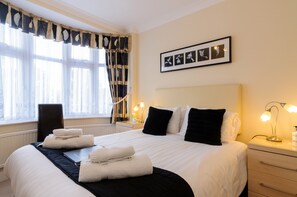A neatly made bed with white and black linens, a bedside table with a lamp, and a window with sheer curtains.