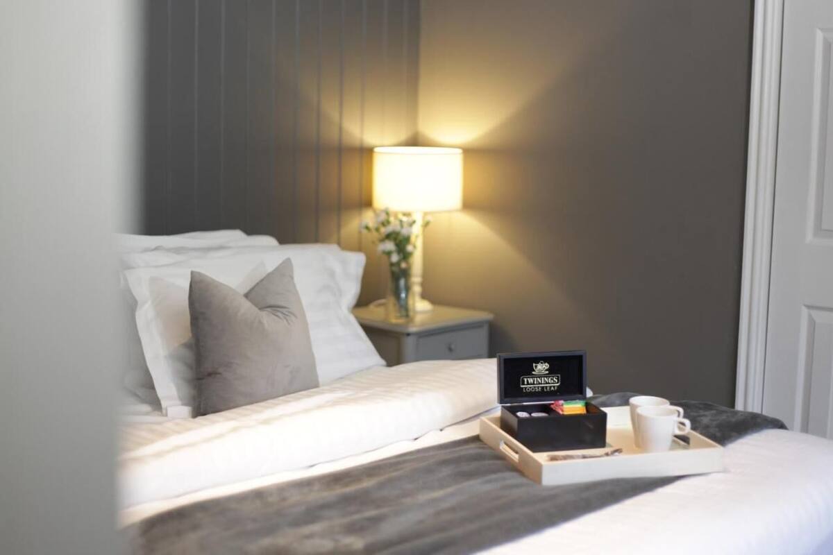 A neatly made bed with pillows, a bedside table with a lamp, and a tray with a box and cups.