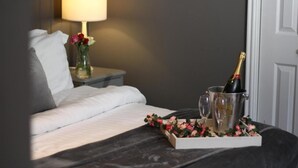 Een netjes opgemaakt bed met witte lakens, een nachtkastje met een lamp, een vaas met bloemen, en een dienblad met een champagnefles en glazen.