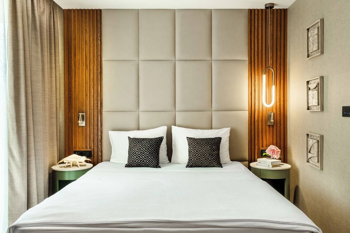 Una habitación de hotel con una cama grande, dos mesitas de noche, un cabecero de madera y una lámpara fijada a la pared.