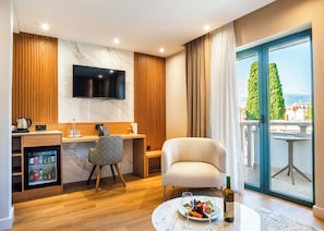 Habitación de hotel moderna con televisor de pantalla plana, minibar, escritorio con silla, zona de comedor con mesa y sillas, y balcón con vistas.