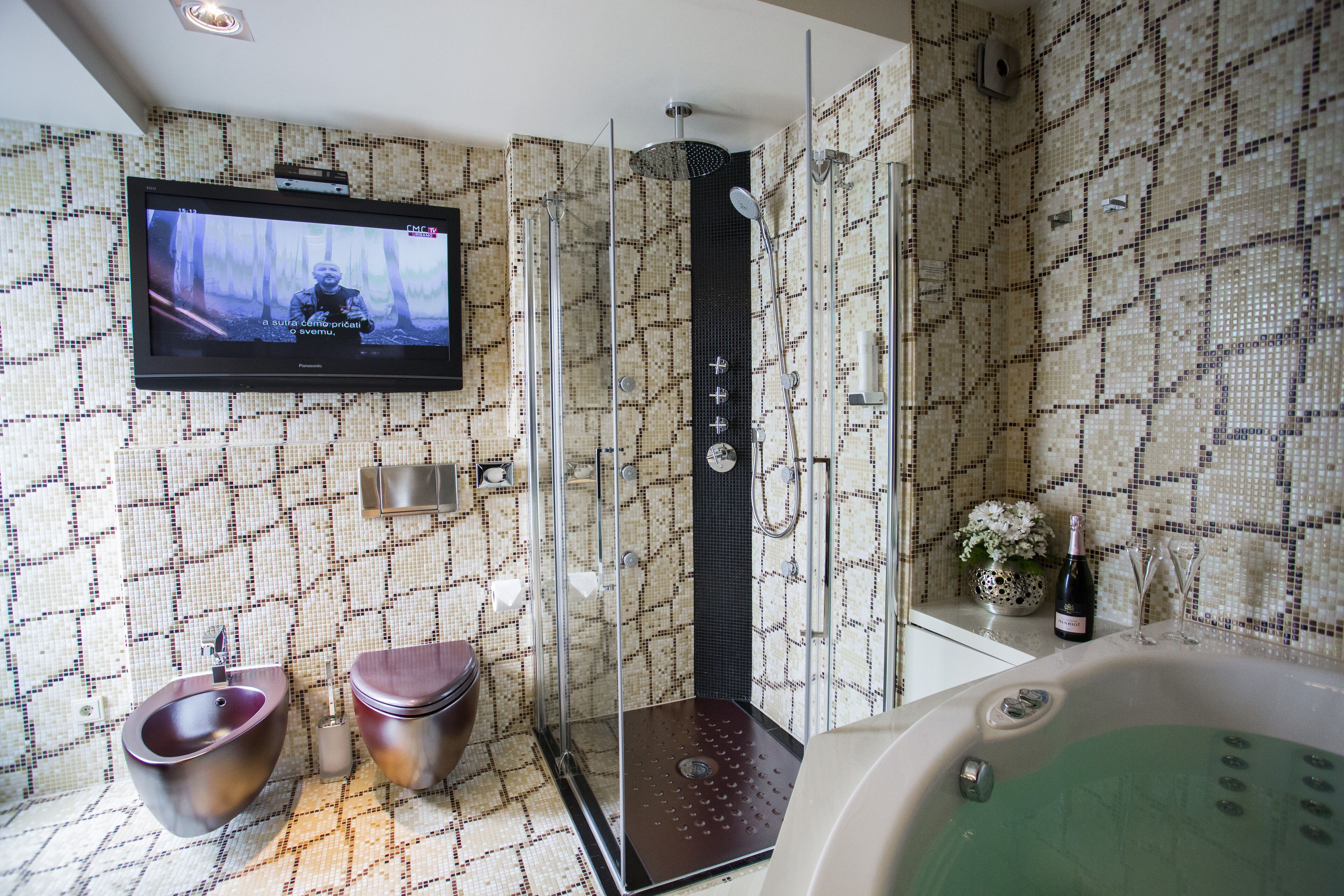 Ein modernes Badezimmer mit einer gläsernen Duschkabine, einem Bidet, einer Toilette, einer Badewanne und einem an der Wand befestigten Fernseher.