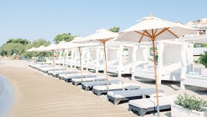 Een hotel aan het strand met rijen witte ligbedden en parasols, een zandpad en een helderblauwe lucht.