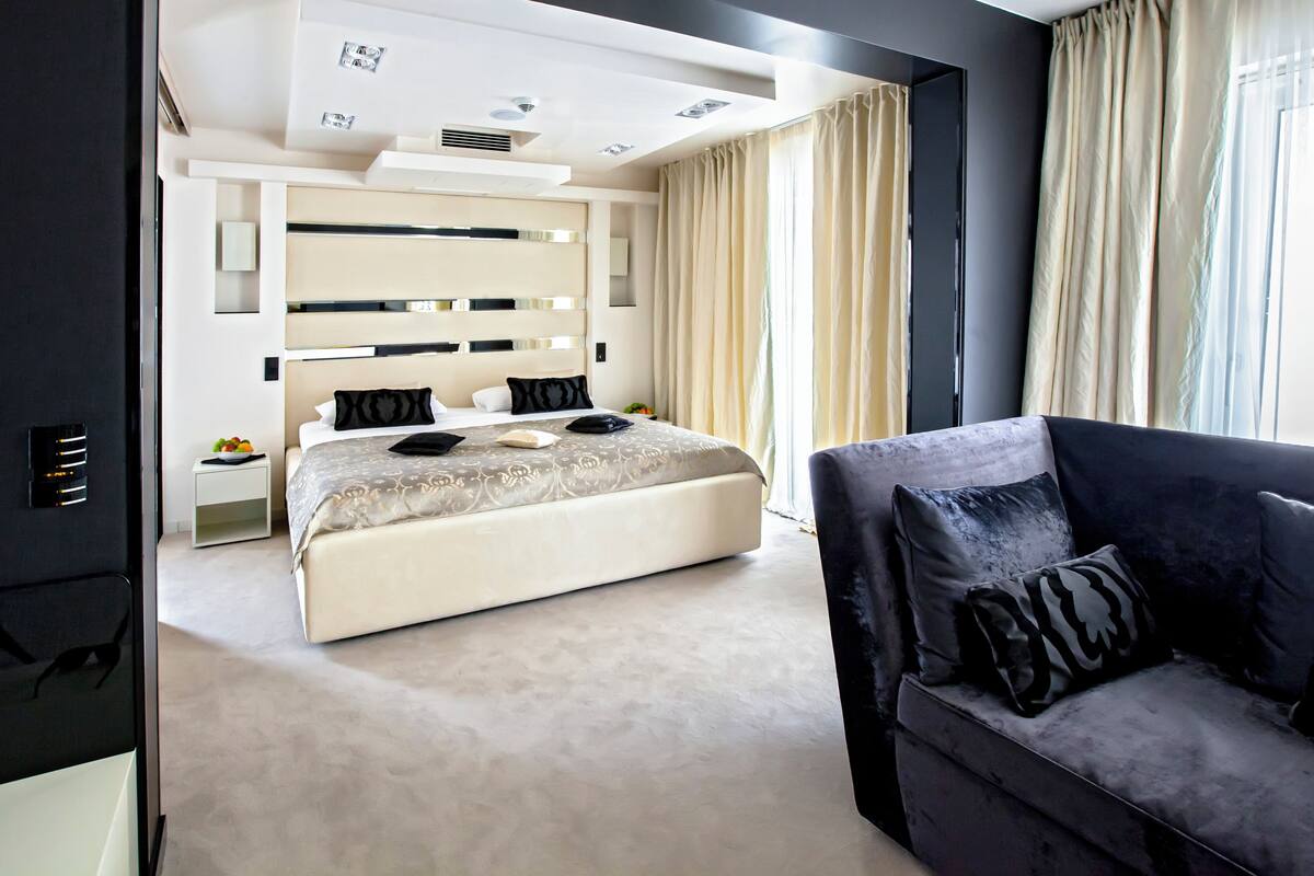Een moderne hotelkamer met een groot bed, een donkere bank en een groot raam met gordijnen.