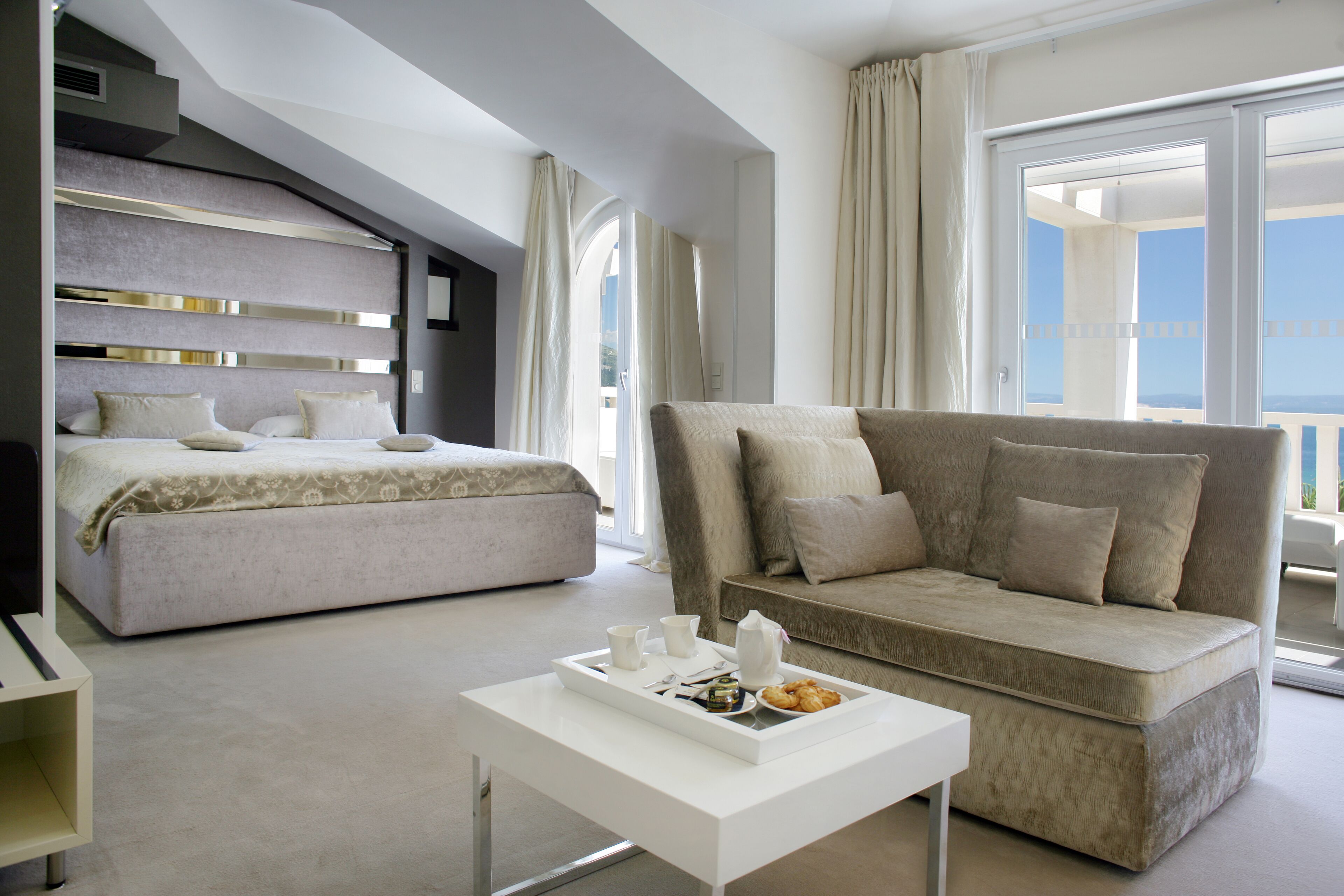 Ein modernes Hotelzimmer mit einem großen Bett, einer Couch, einem Couchtisch mit einem Tablett mit Frühstücksangeboten und einem Balkon mit Blick.