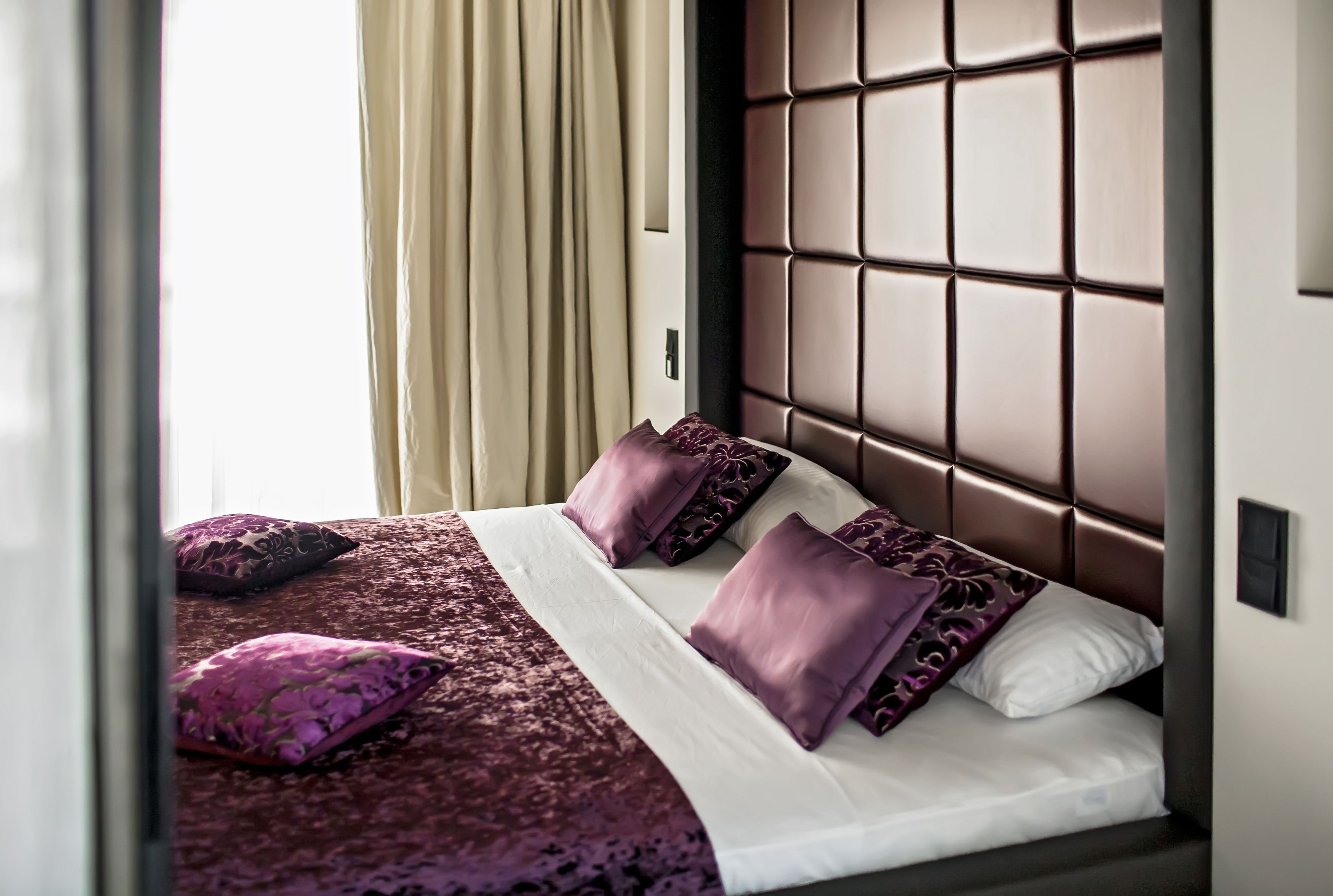 A hotel room with a large bed, a headboard with a grid pattern, and a window with curtains.