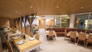 A restaurant with wooden ceiling and walls, tables with yellow tablecloths, and a large window offering a view. - Hotel Majorka (Andalo)