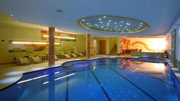 A modern indoor swimming pool with a curved design, surrounded by lounge chairs and a Coca-Cola branded wall. - Hotel Majorka (Andalo)