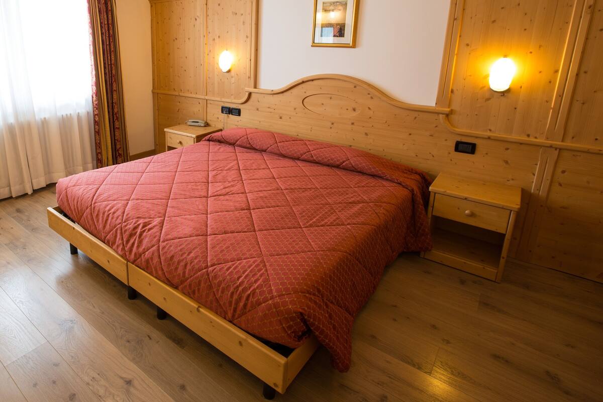 A wooden bed with a quilted red bedspread, a nightstand with a lamp, and a wooden wardrobe.