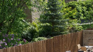 An outdoor patio with multiple round tables and wicker chairs, surrounded by a wooden fence and lush greenery.