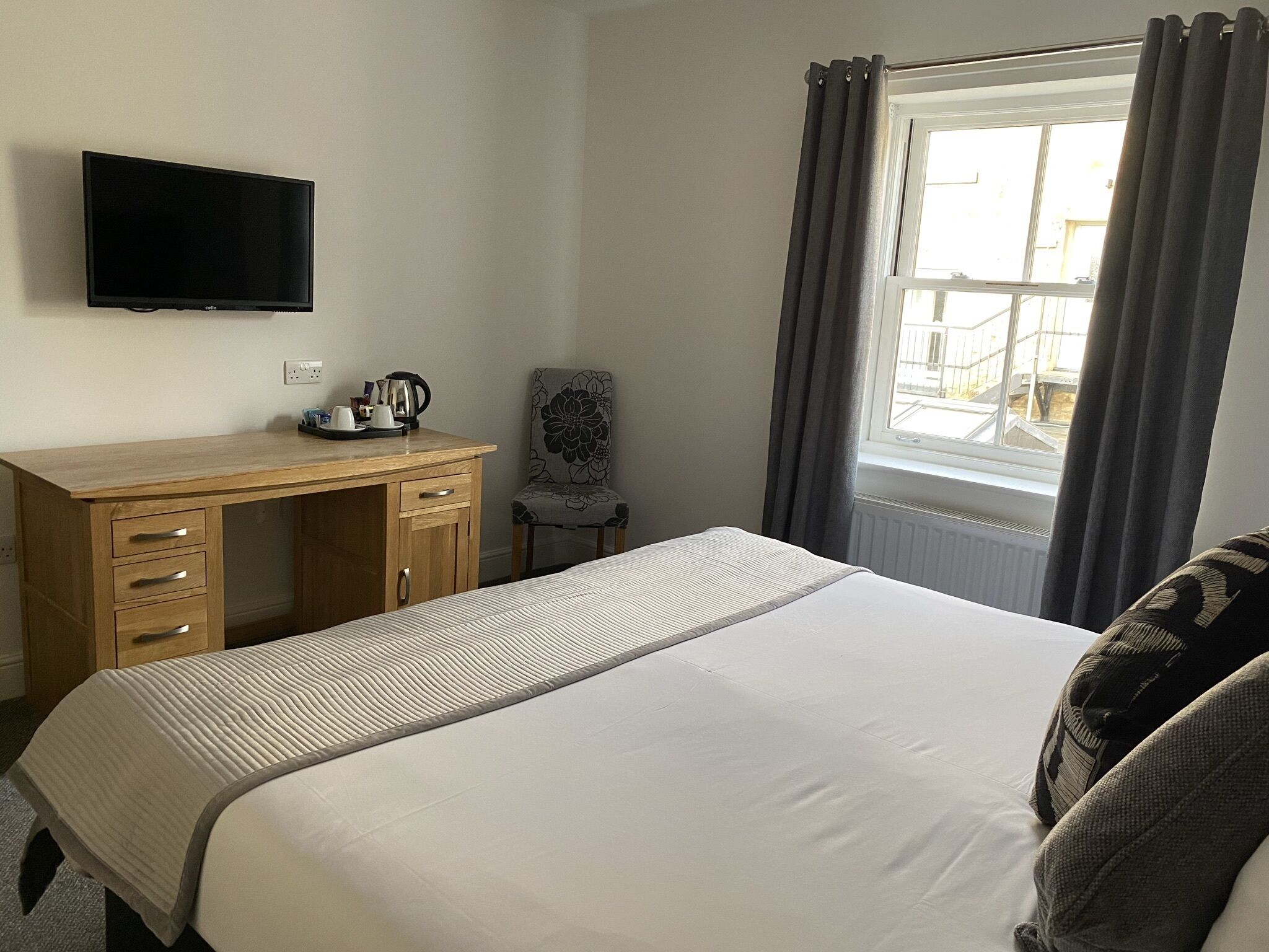 Ein Hotelzimmer mit einem Holztisch, einem Flachbildfernseher, einem Bett mit gestreifter Bettdecke, einem Sessel und einem Fenster mit Vorhängen.