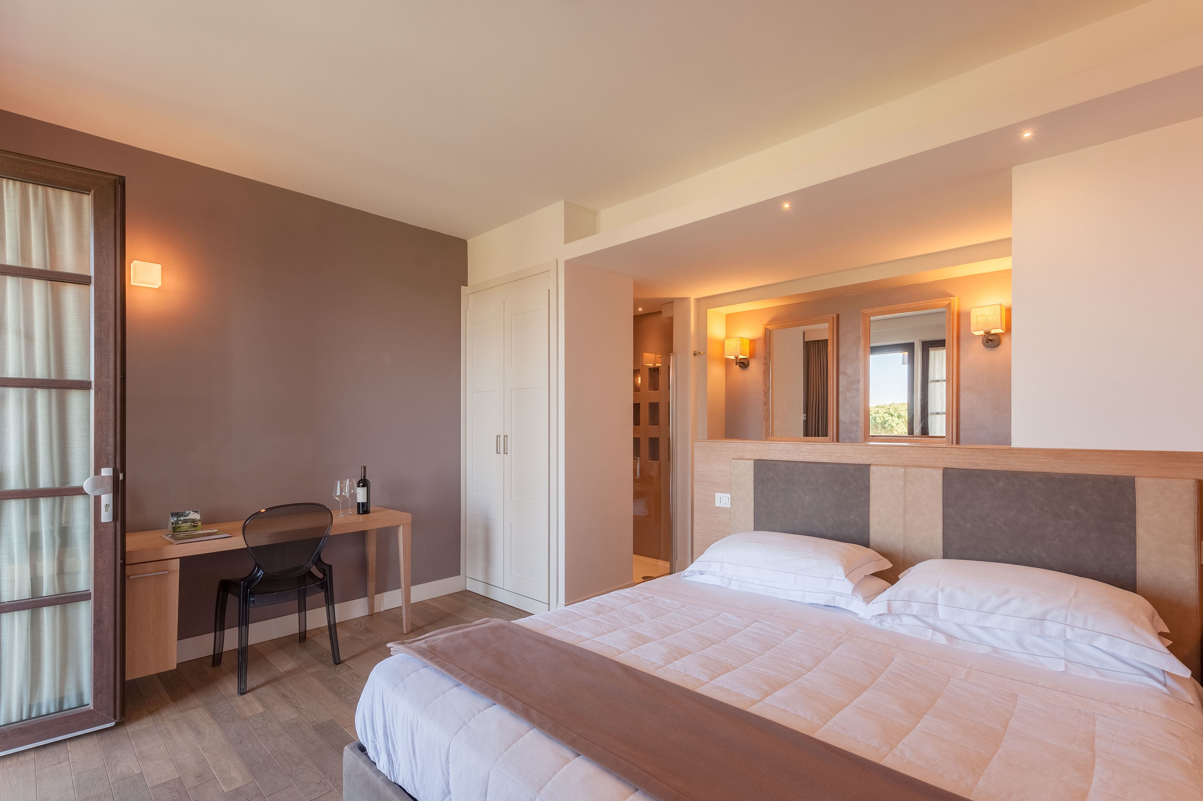 A hotel room with a bed, a desk, and a chair. There is a wardrobe, a mirror, and a window.