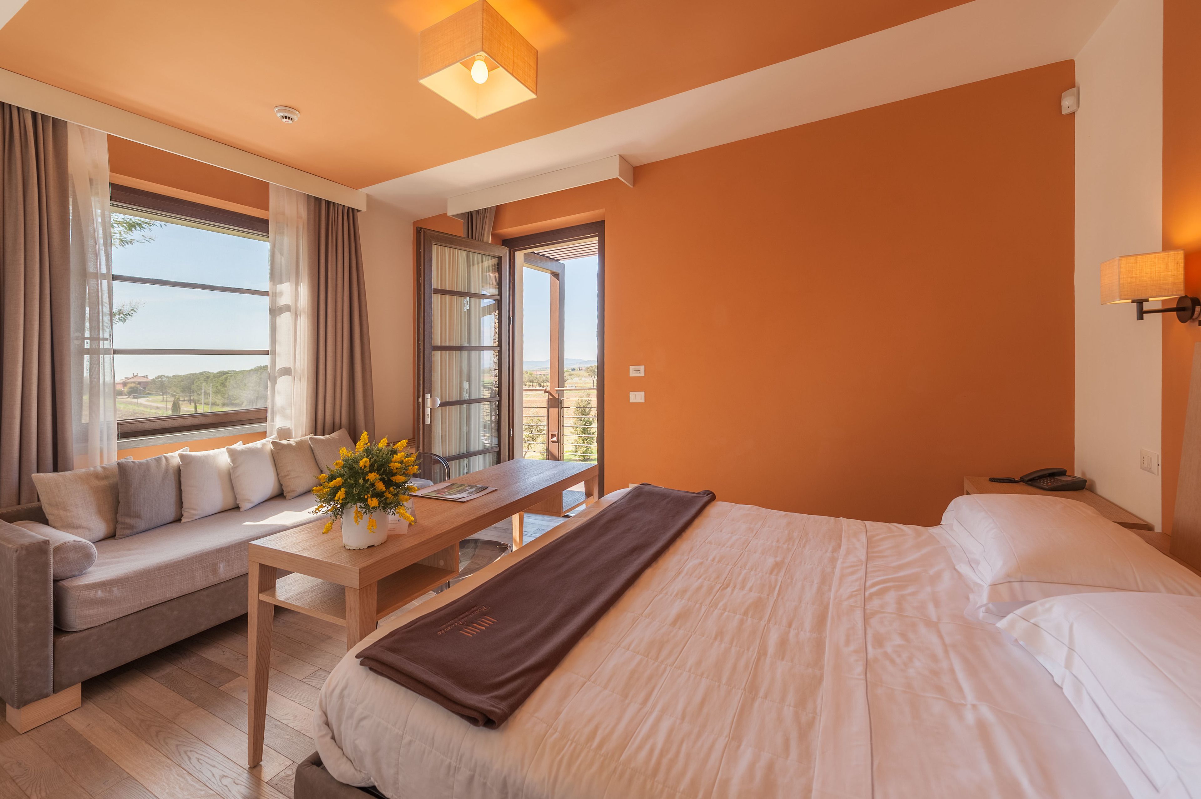 A modern hotel room with a large bed, a sofa, a wooden table with a vase of flowers, and a view of the outdoors through a large window.