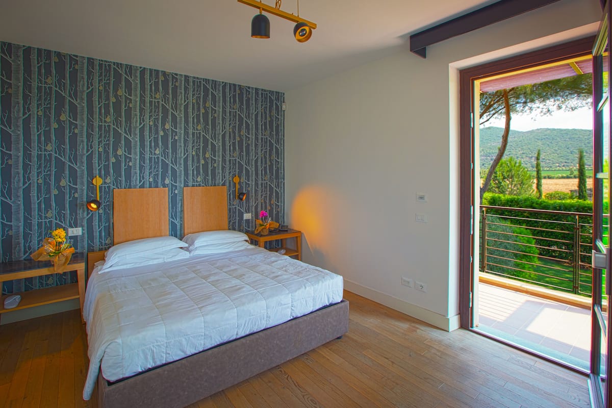 Un dormitorio con una cama grande, una mesita de noche con una lámpara y vistas a un balcón con vegetación.