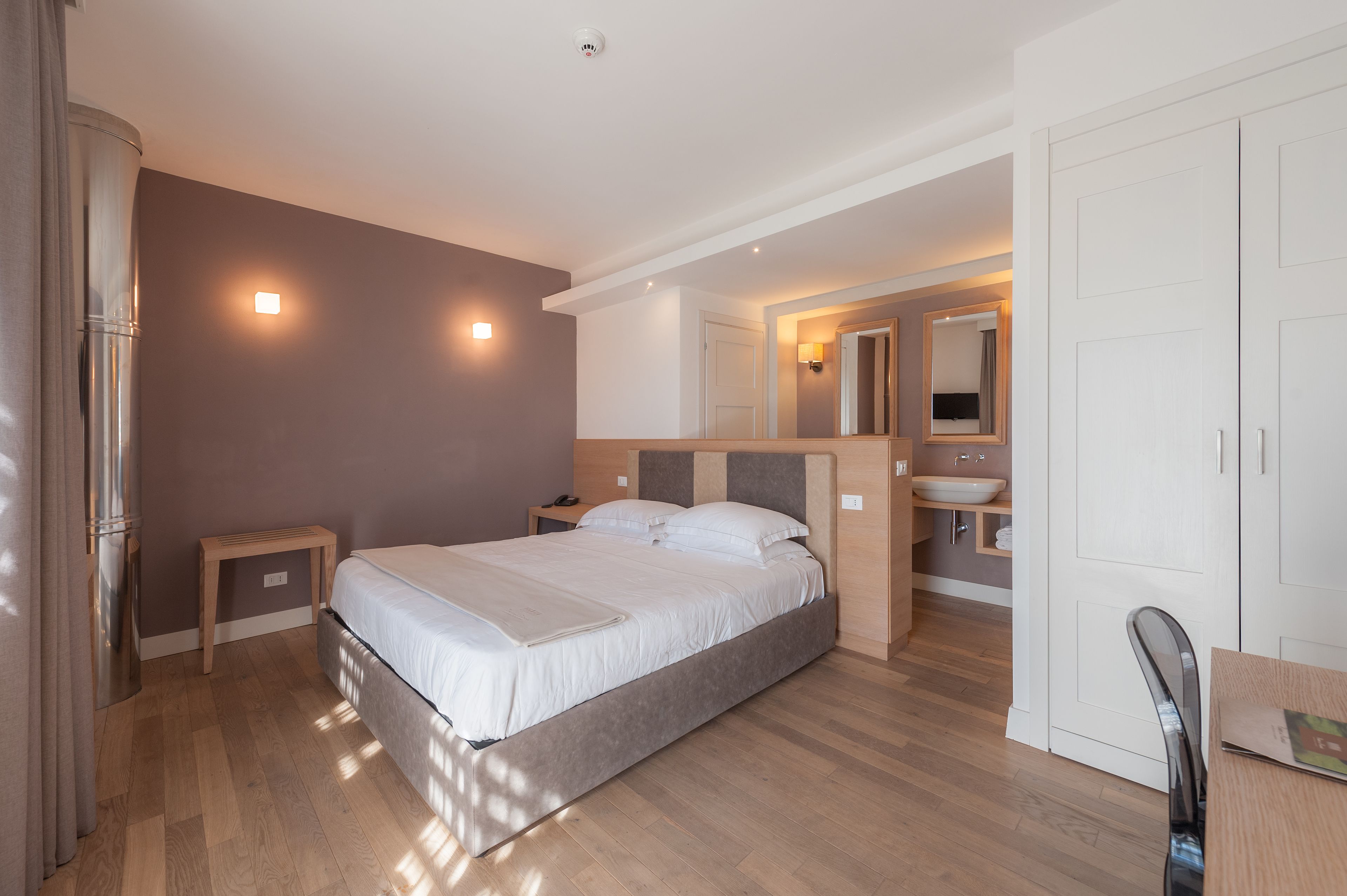 Un dormitorio moderno con una cama grande, mesitas de noche, una mesita auxiliar, una silla de madera, un escritorio y un baño con lavamanos y espejo.