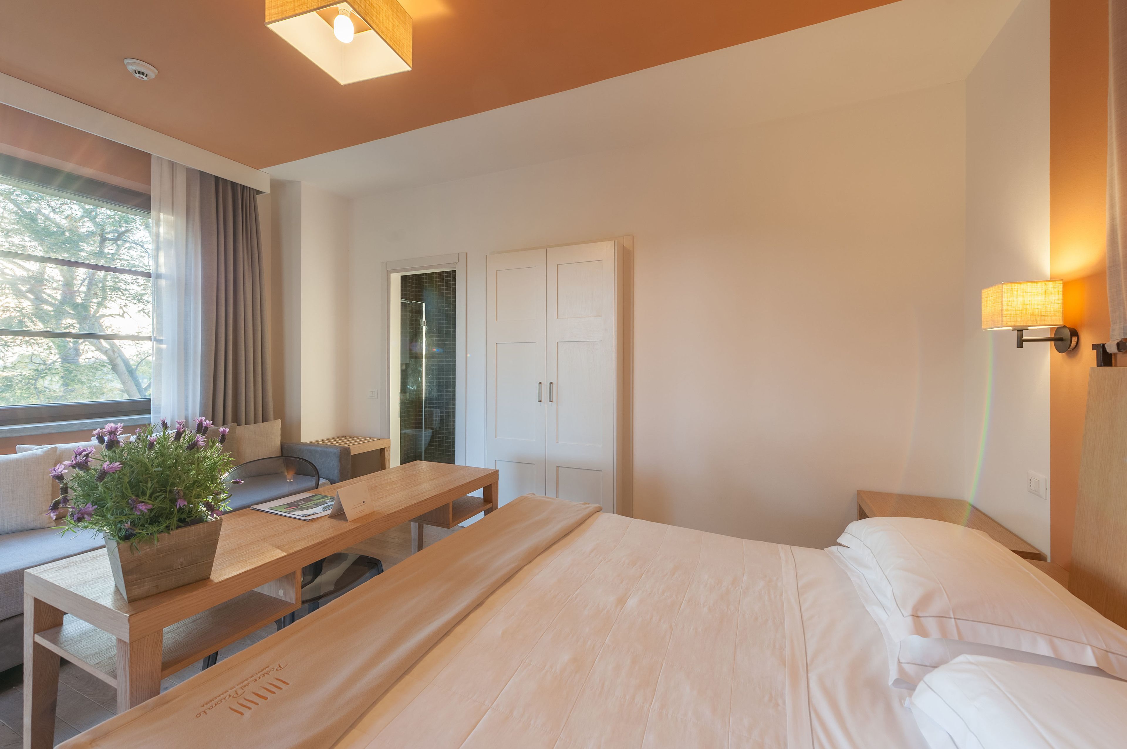A modern hotel room with a bed, a wooden desk, a seating area with a bench, a wardrobe, and a window with curtains.