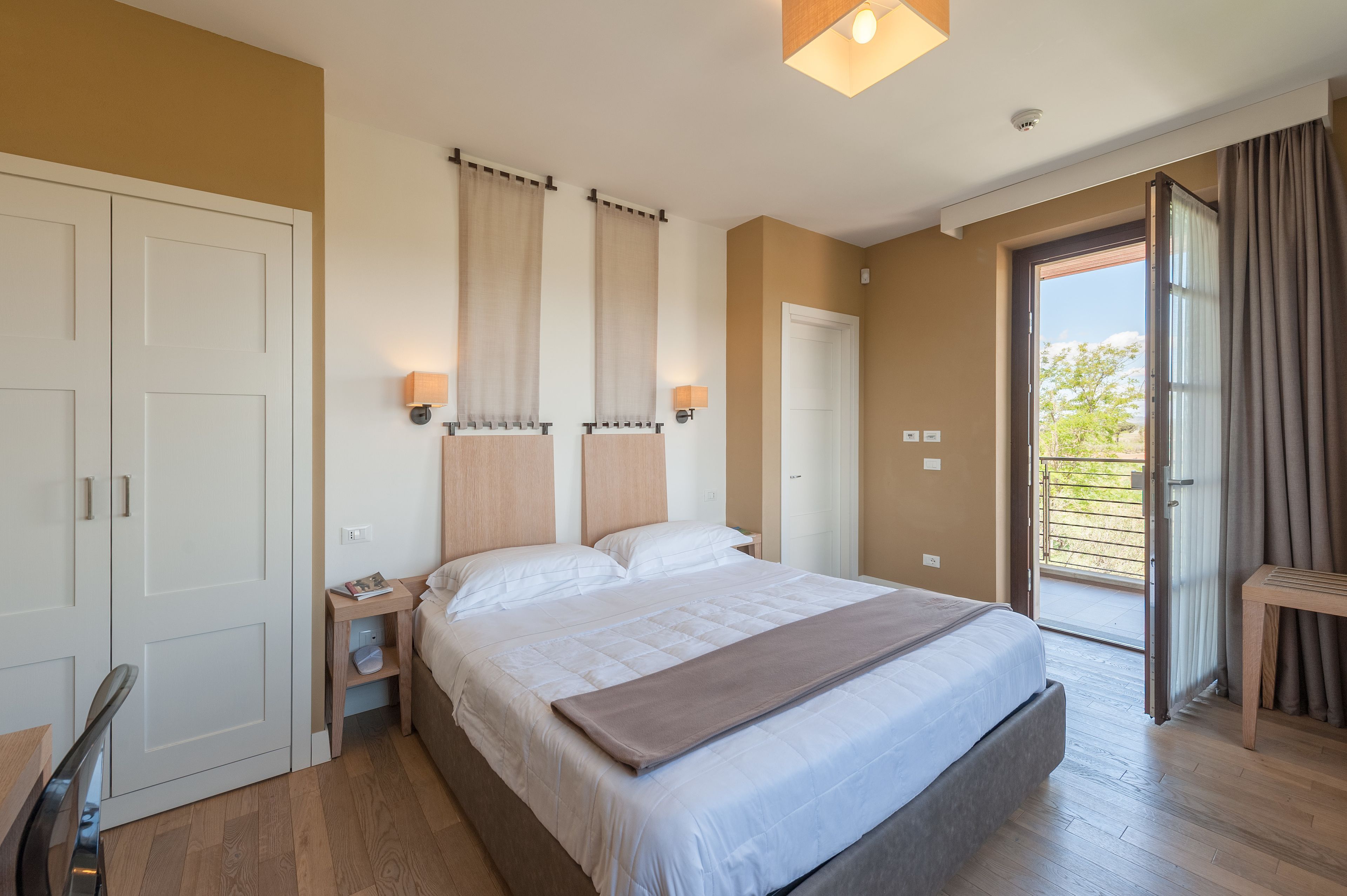 A bedroom with a large bed, two bedside tables, a nightstand, a sliding glass door to a balcony, and a wooden chair.